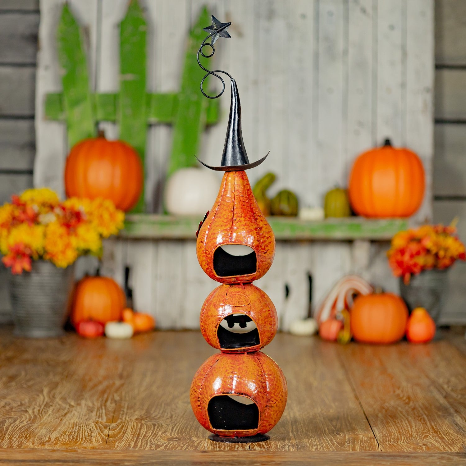 A decorative stacked pumpkin sculpture in orange with black carved facial features, topped with a black witch hat and star, displayed on a wooden surface with pumpkins and fall flowers in the background.