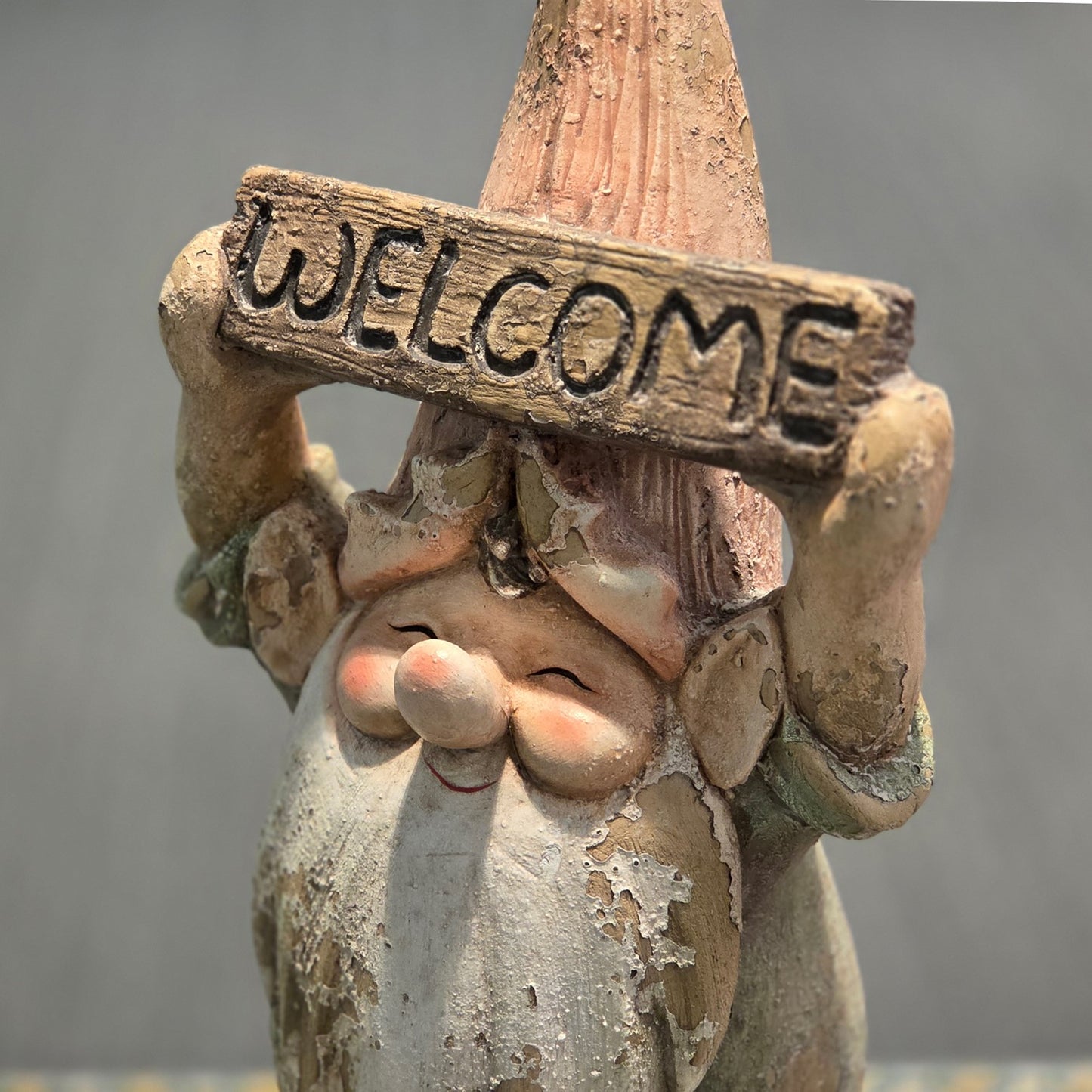A close-up of a decorative gnome figurine holding a wooden sign that reads 'WELCOME' in raised letters. The gnome has a weathered, rustic finish with peeling paint-like texture, a pointed hat, and a long beard, set against a soft gray background.