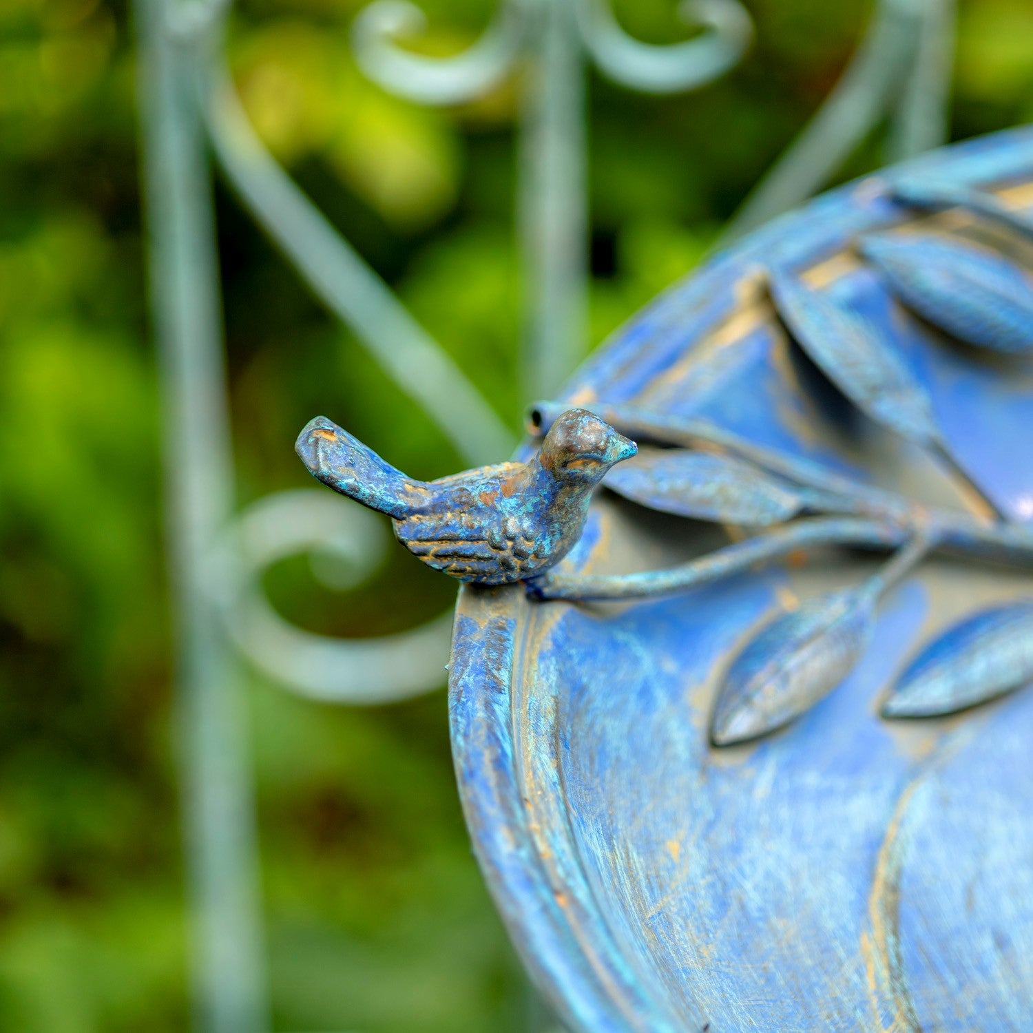 A close-up shot of a small bird figurine perched on a blue metal garden bench with leaf designs, set against a blurred green garden background.