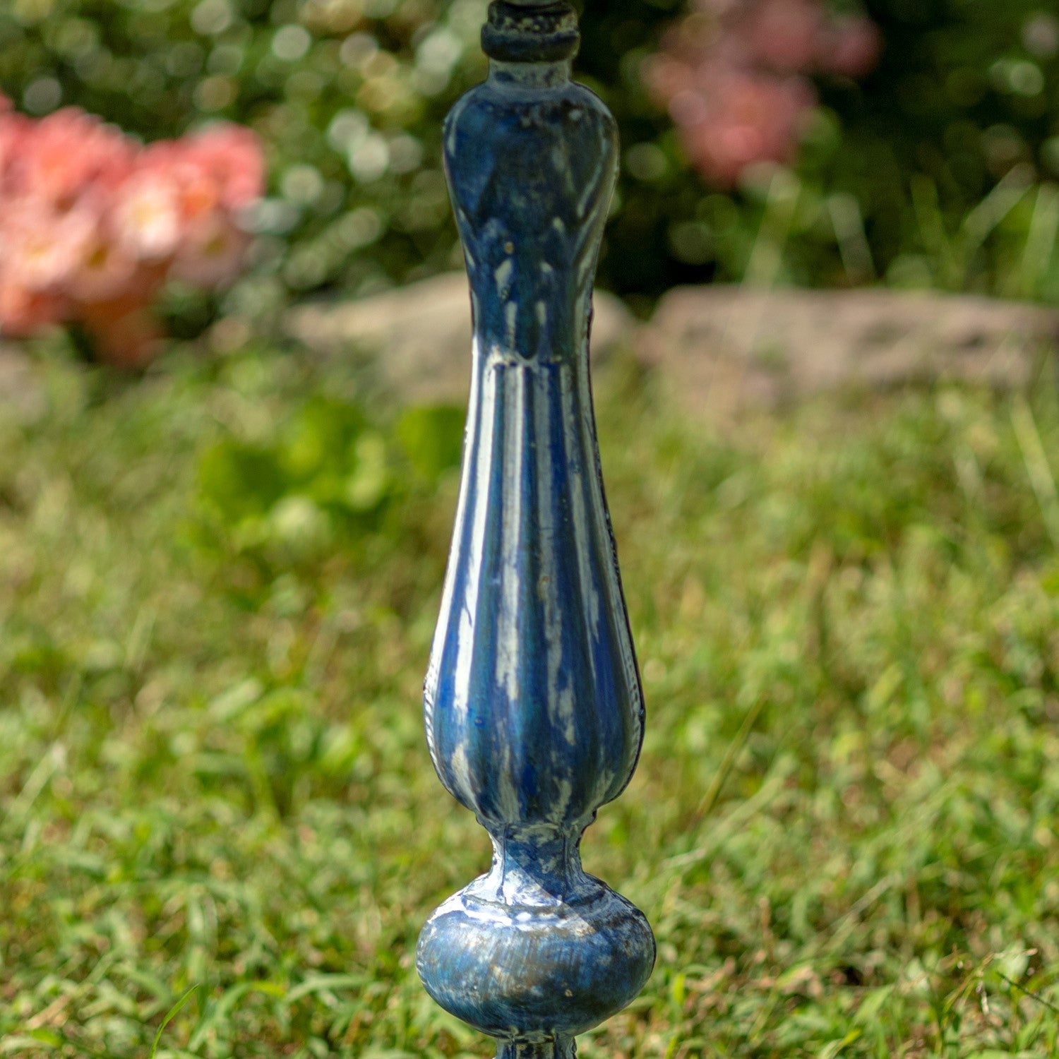 A blue and white striped decorative garden stake or finial standing upright in a grassy outdoor setting with blurred greenery and flowers in the background.