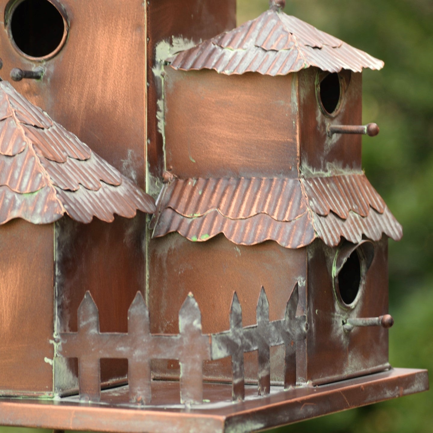 A close-up of a decorative copper birdhouse with tiered shingled roofs, a picket fence detail, and multiple circular entry holes, shown against a blurred green outdoor background.