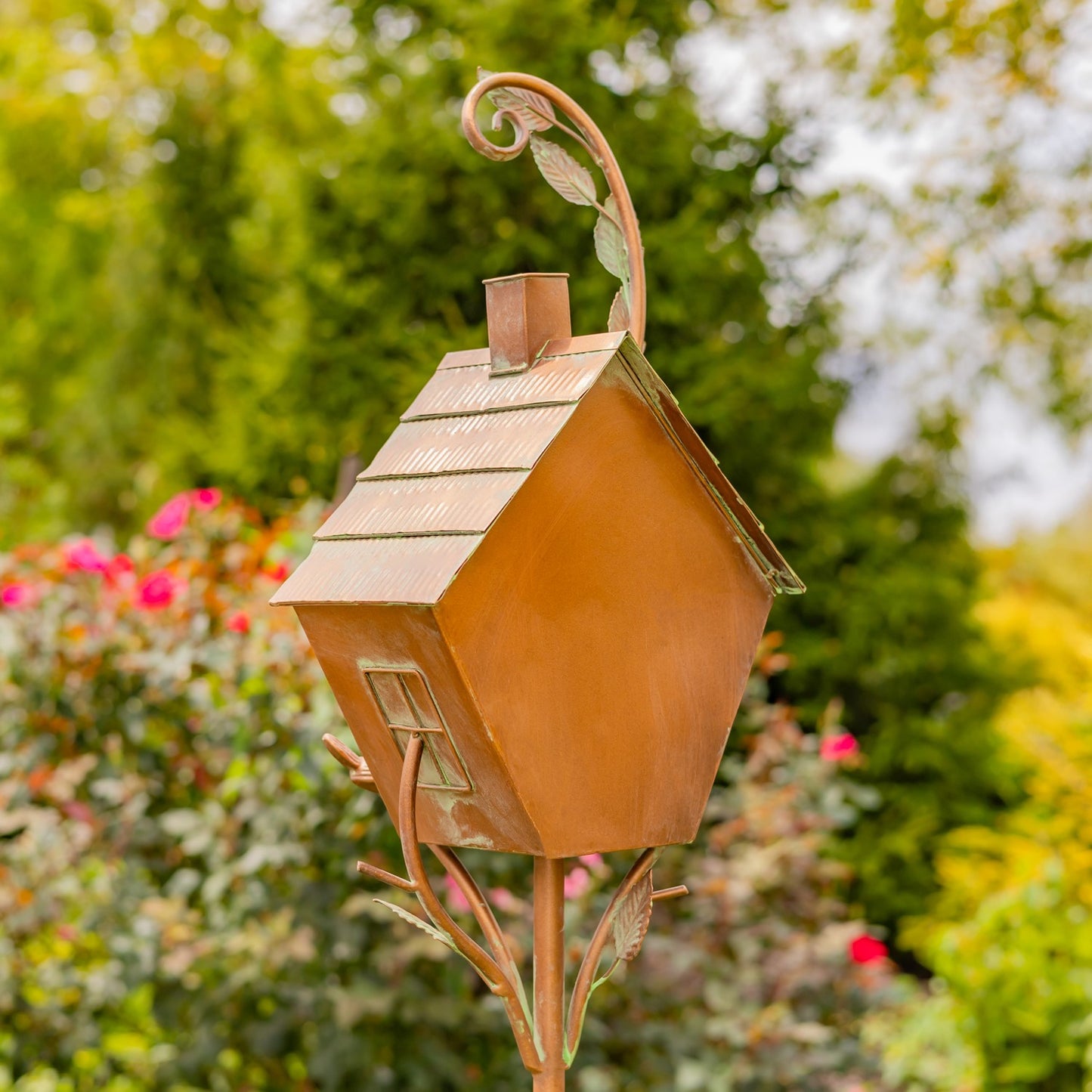 Decorative copper birdhouse with a small bird on top, set against a garden background with flowers and trees