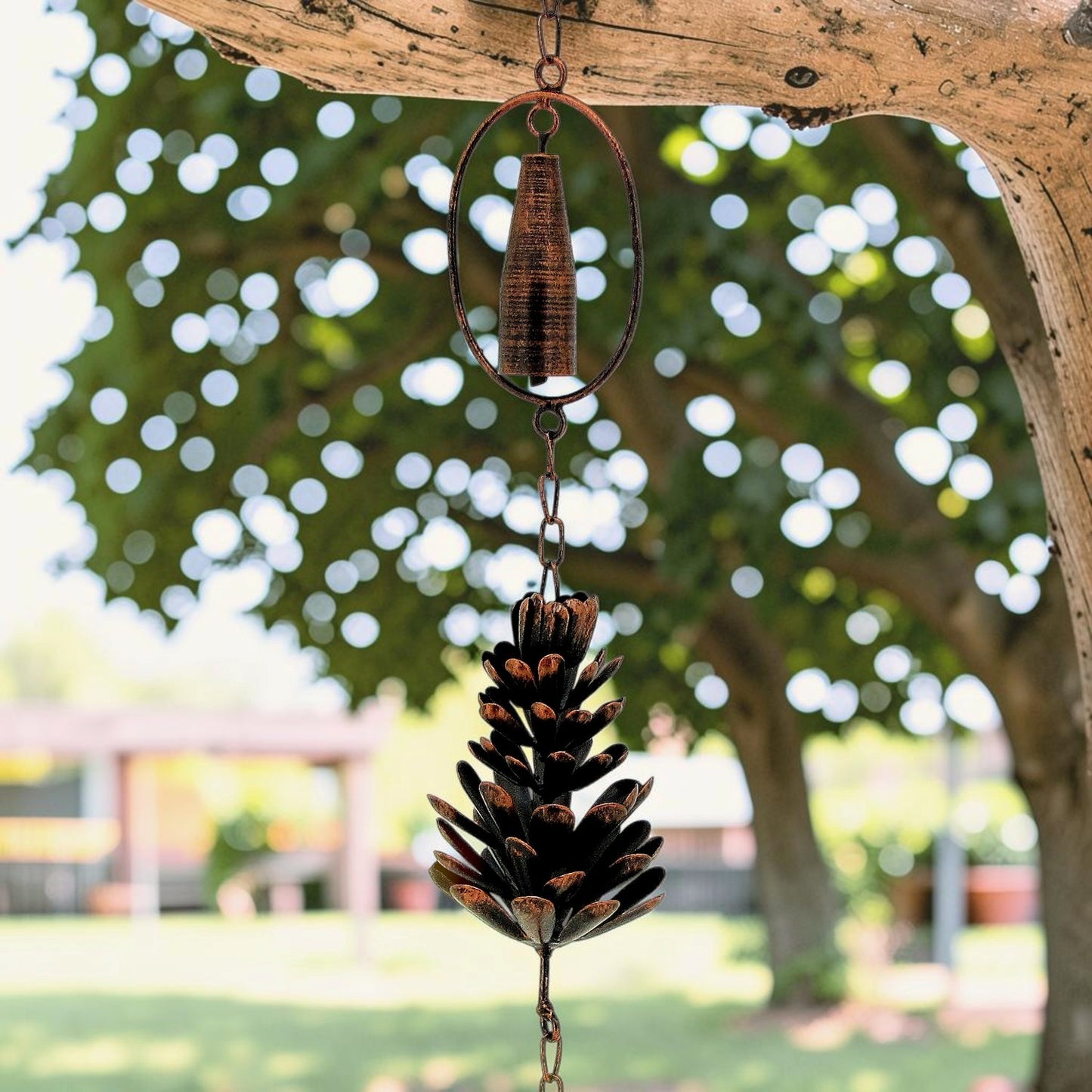 A rustic metal wind spinner ornament featuring a pinecone-shaped centerpiece and a hanging ring, suspended from a tree branch in an outdoor garden setting.