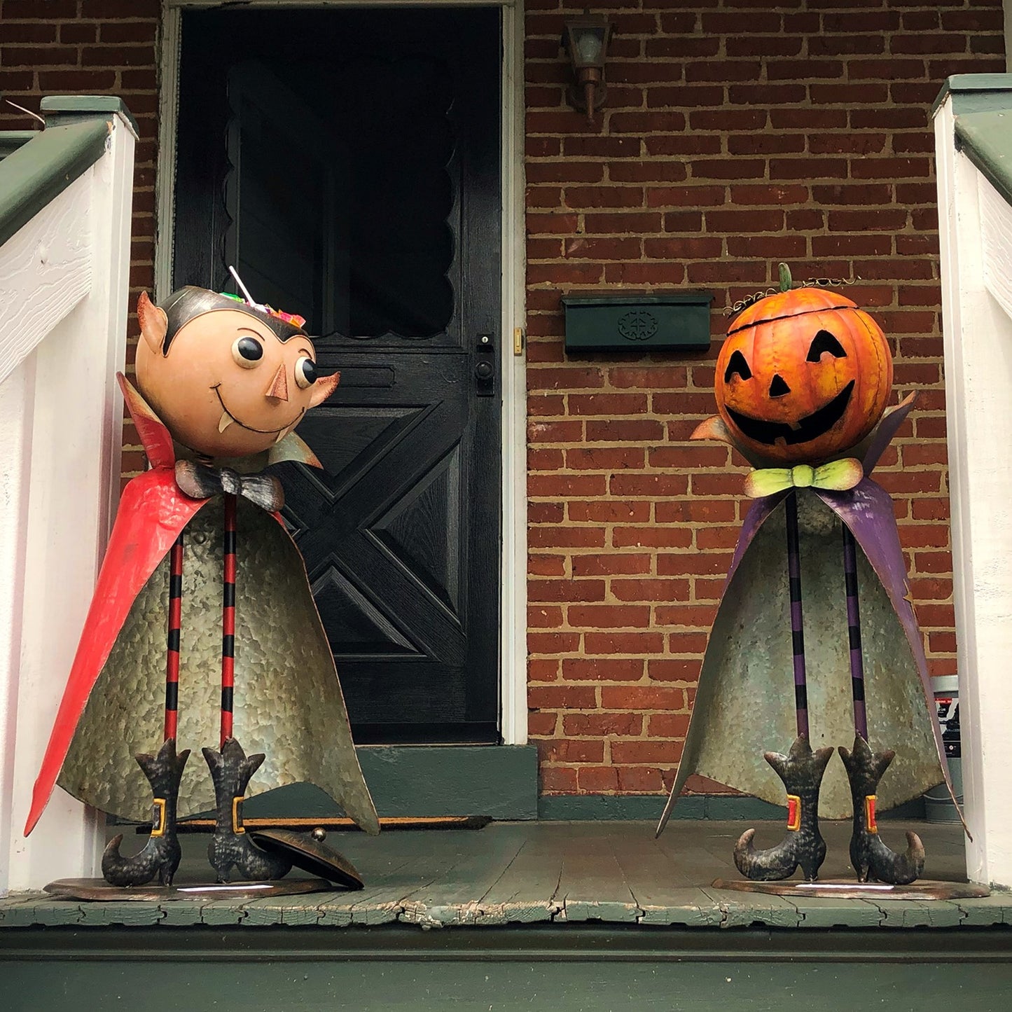 Two Halloween yard decorations on a front porch: a vampire figure with a red cape and a jack-o'-lantern figure with a purple cape, set against a brick house door.