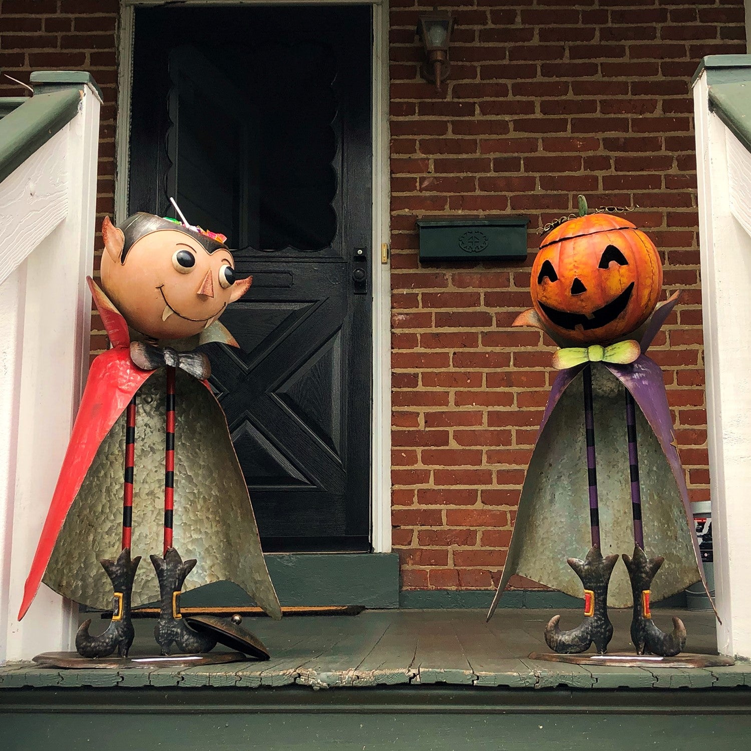 Two Halloween yard decorations on a front porch: a vampire figure with a red cape and a jack-o'-lantern figure with a purple cape, set against a brick house door.