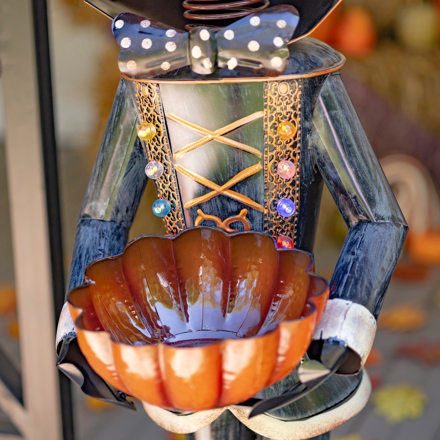 A decorative metal garden statue shaped like a nutcracker holding a glossy orange pumpkin bowl, photographed close up with a blurred outdoor background.