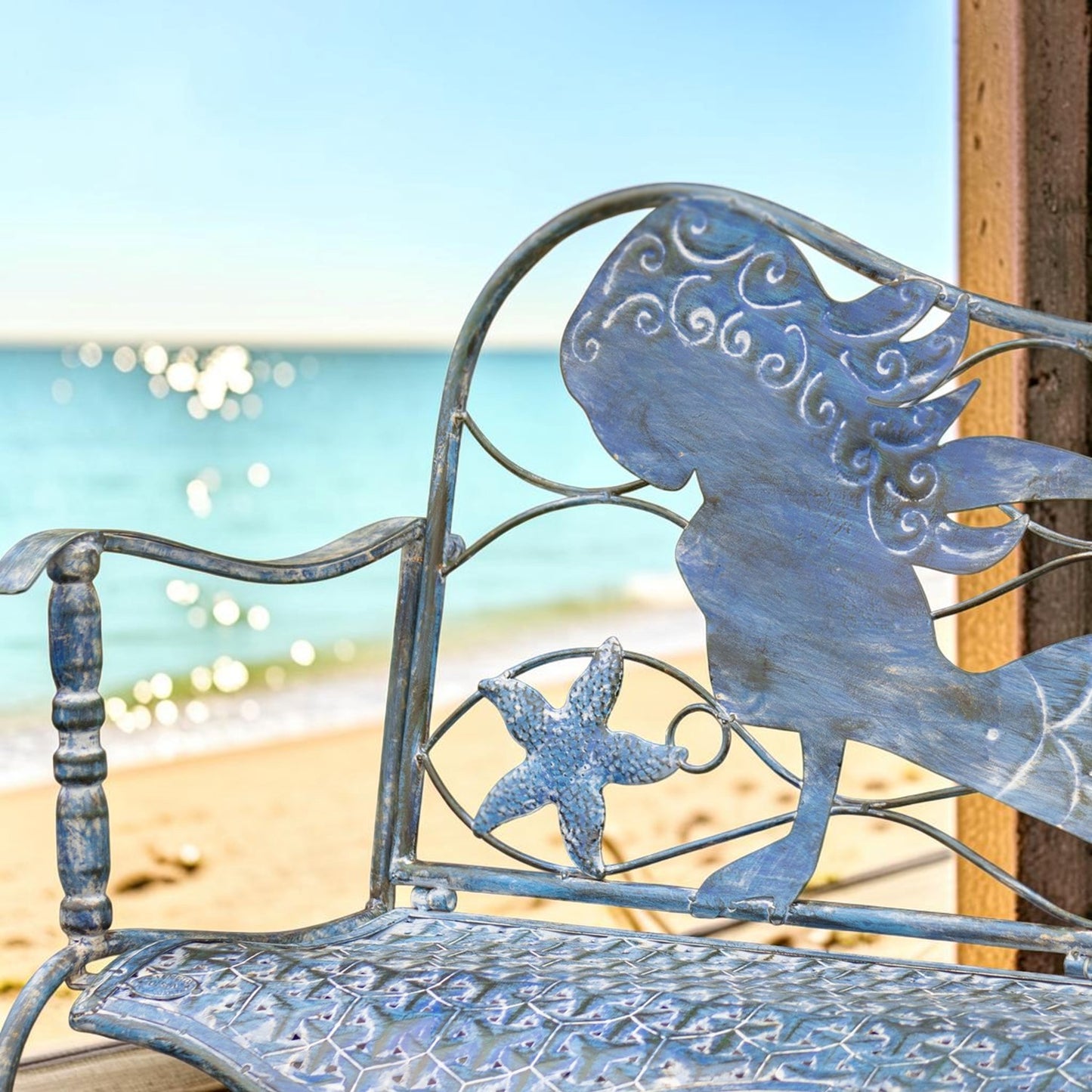 A blue metal beach bench with a mermaid silhouette and starfish design, photographed outdoors with a blurred ocean and sandy beach background.