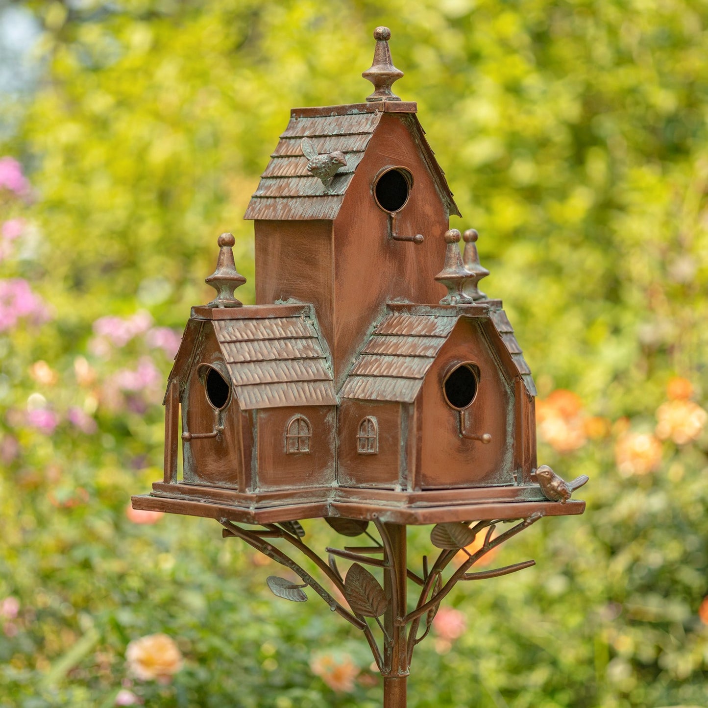 A decorative multi-tiered birdhouse with a rustic copper-brown finish and weathered green patina details, mounted on a metal stand against a blurred green garden background.