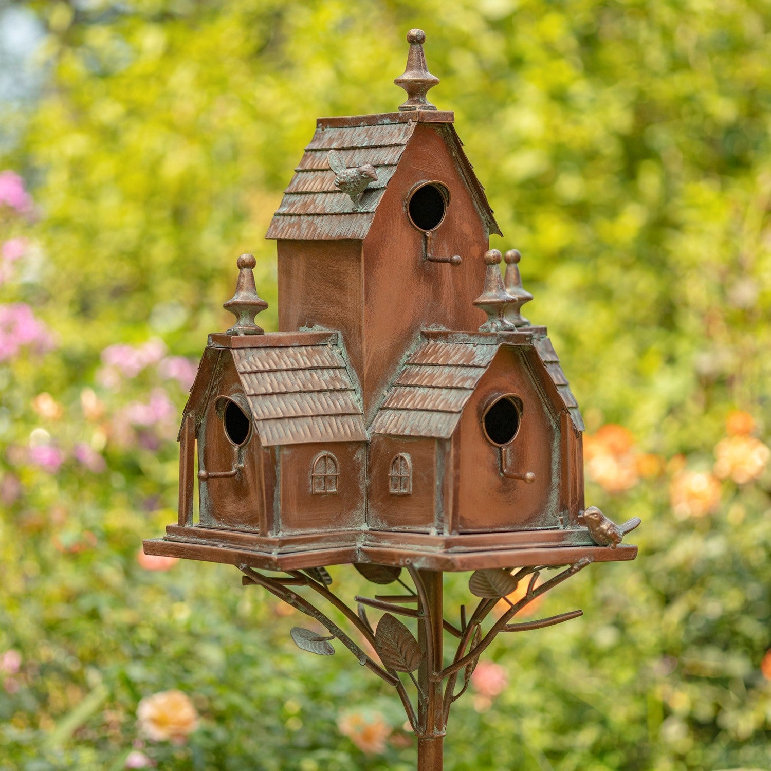 A decorative multi-tiered birdhouse with a rustic copper-brown finish and weathered green patina details, mounted on a metal stand against a blurred green garden background.
