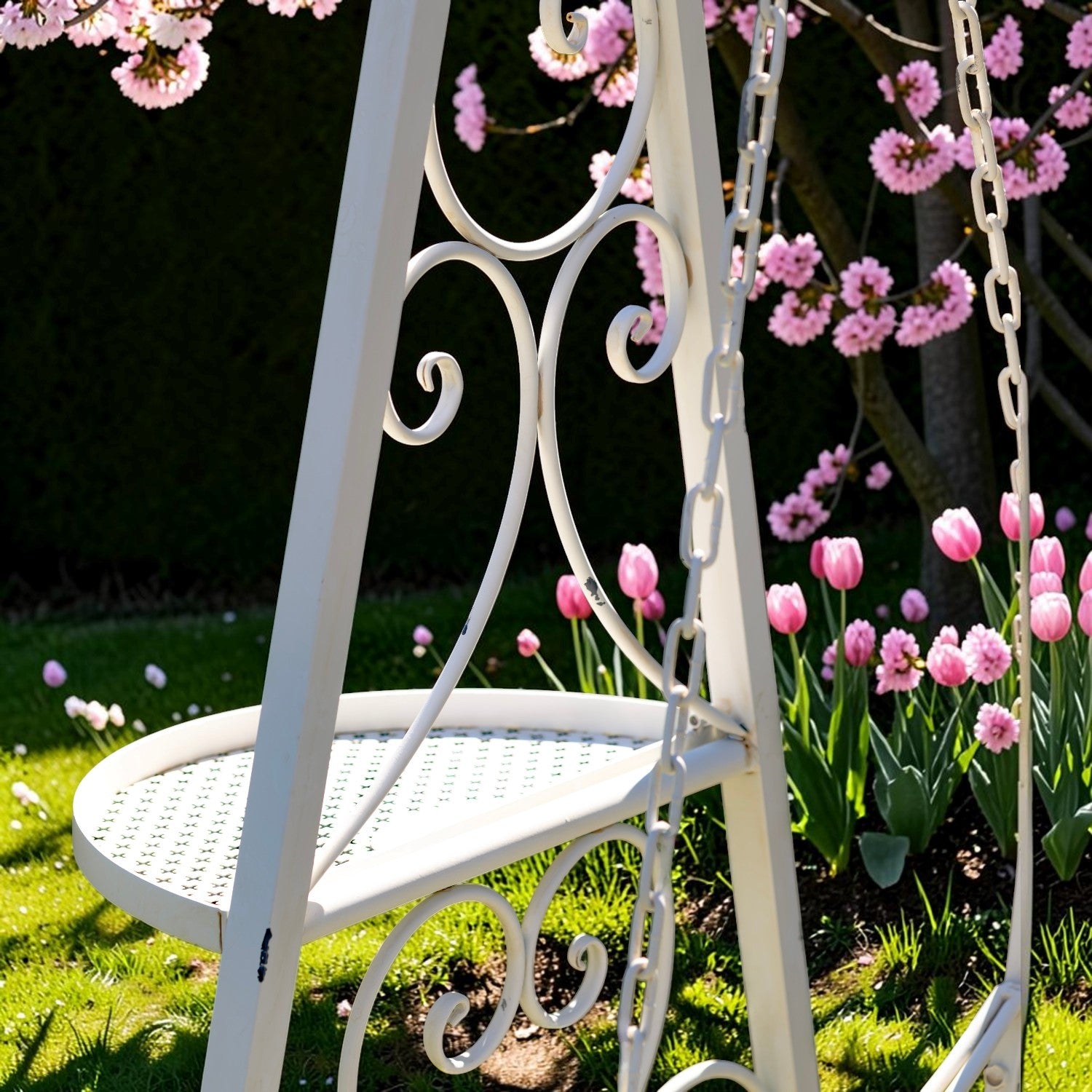 A white wrought-iron garden swing chair with decorative scrollwork and hanging chains, set in a grassy garden with pink flowers in bloom.