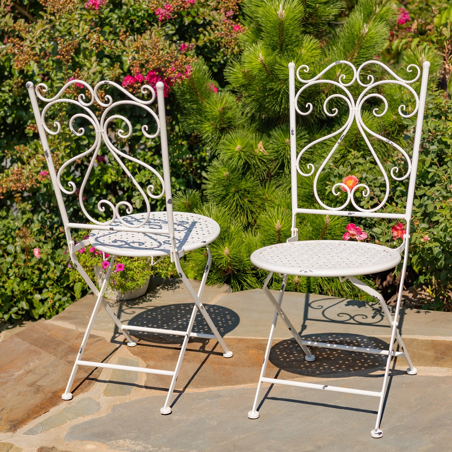 Two white wrought iron folding garden chairs with decorative scrollwork backs, shown on a stone patio in front of green garden foliage and pink flowers.