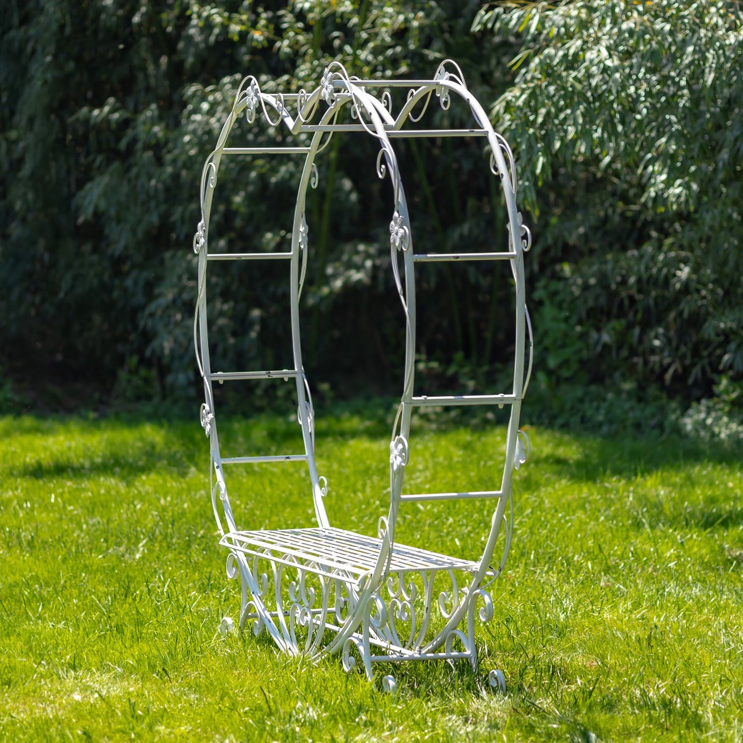 A white wrought iron garden bench with an arched trellis design and ladder-style side supports, placed on a green grassy lawn with leafy trees in the background.