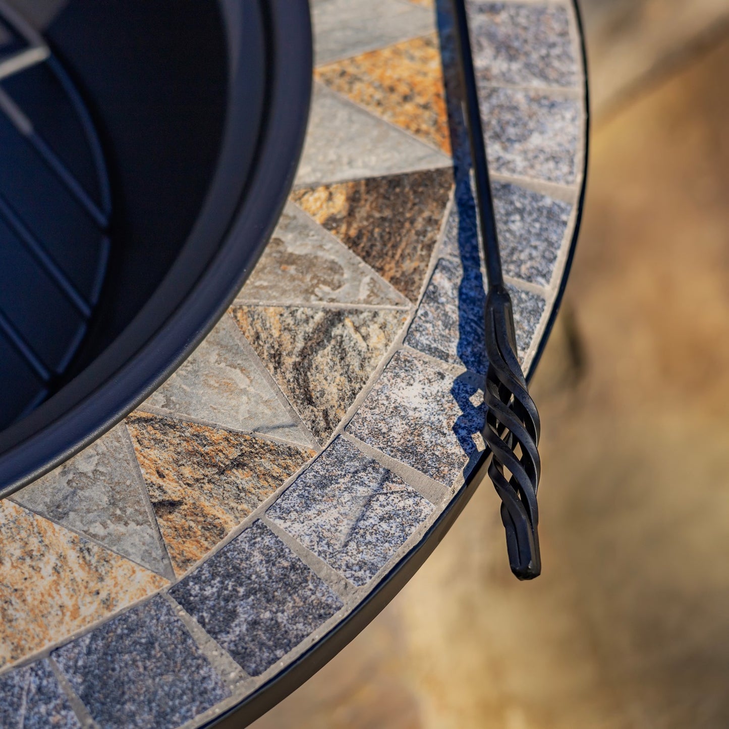 Close-up of a round outdoor fire pit table with a mosaic stone tile pattern in gray, beige, and brown, featuring a black metal rim and a black fire poker.