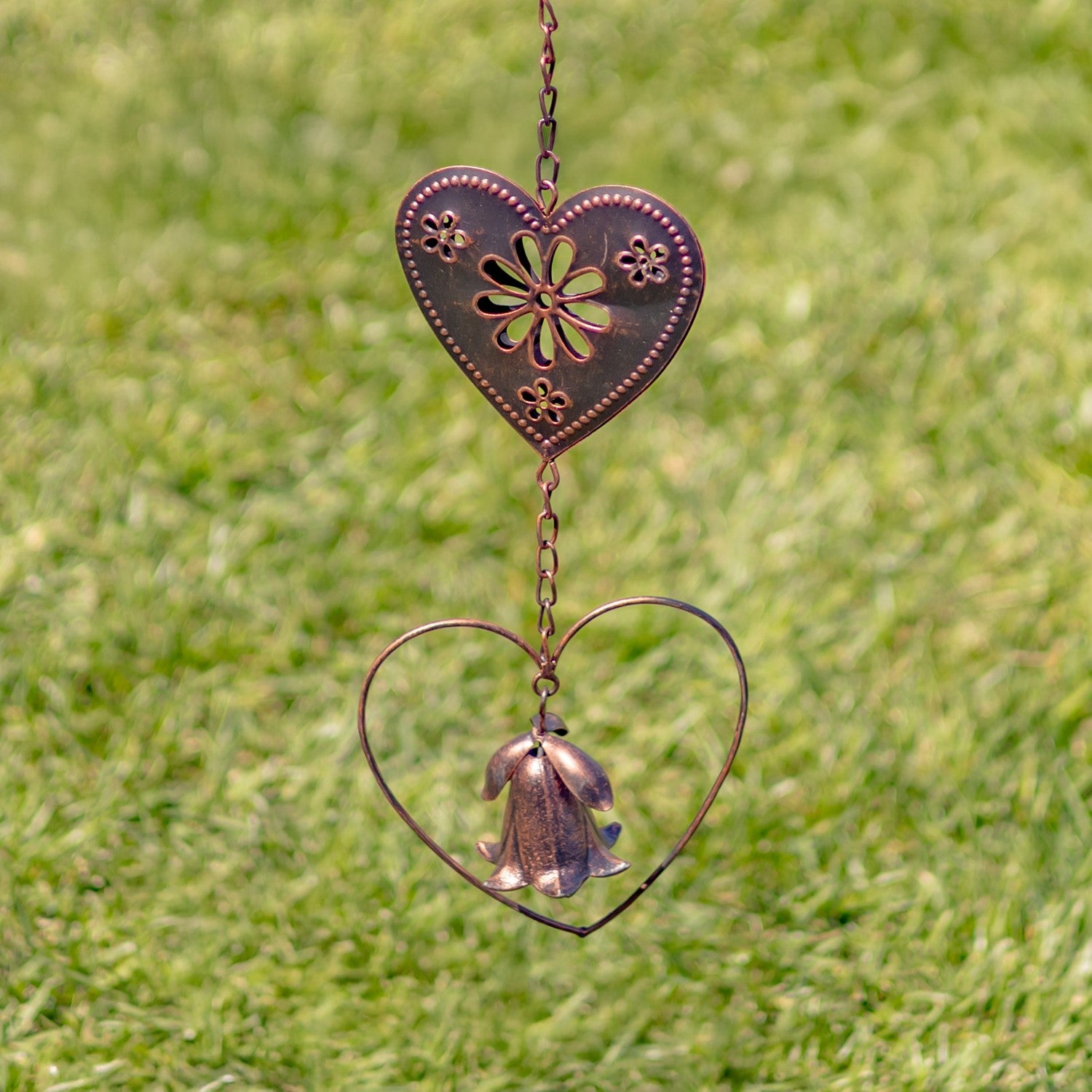 A copper-toned metal garden wind spinner featuring a bell-shaped flower suspended inside a heart-shaped wire frame, set against a blurred green grass background.