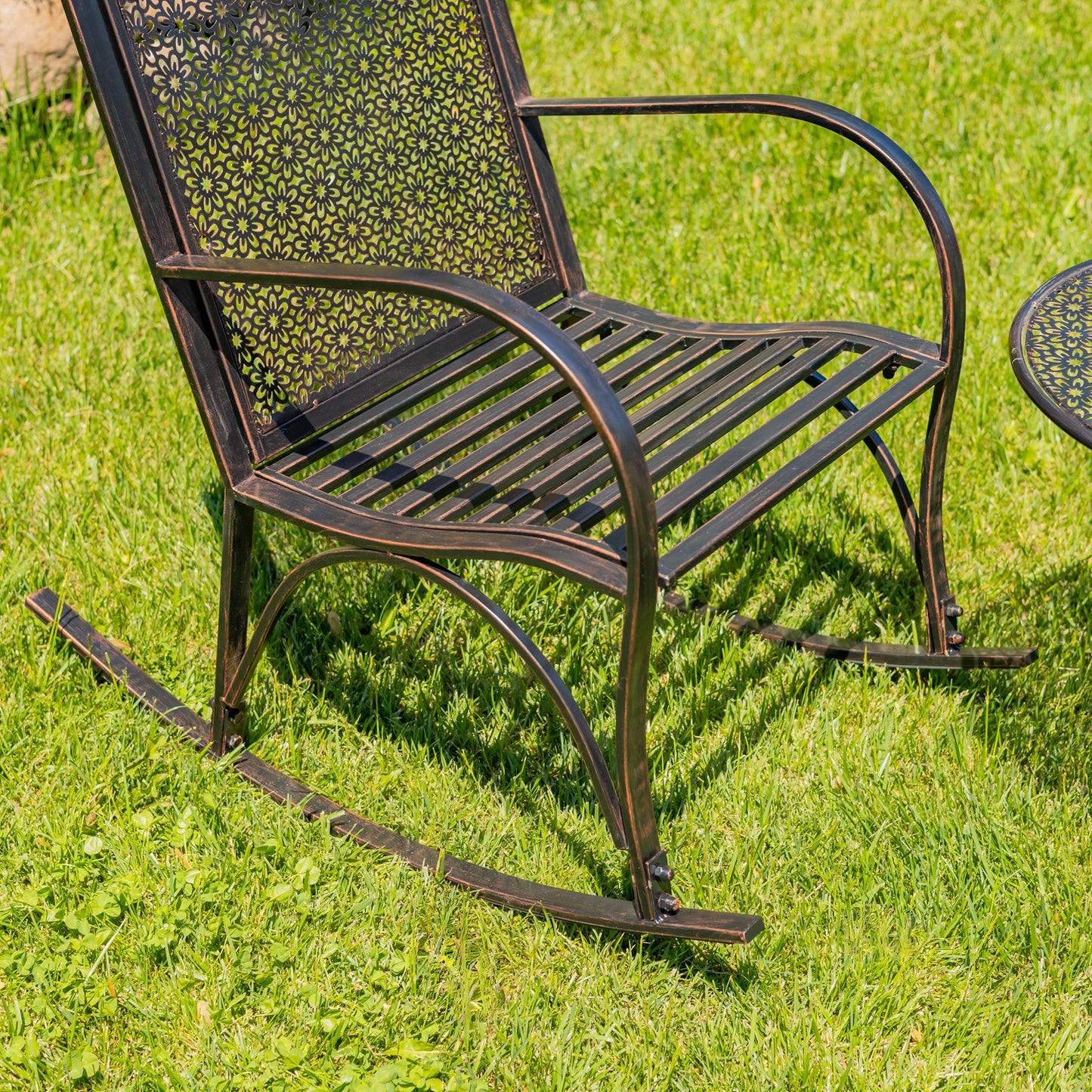 A dark bronze metal rocking chair with a perforated backrest and curved armrests, shown outdoors on a green grass lawn.
