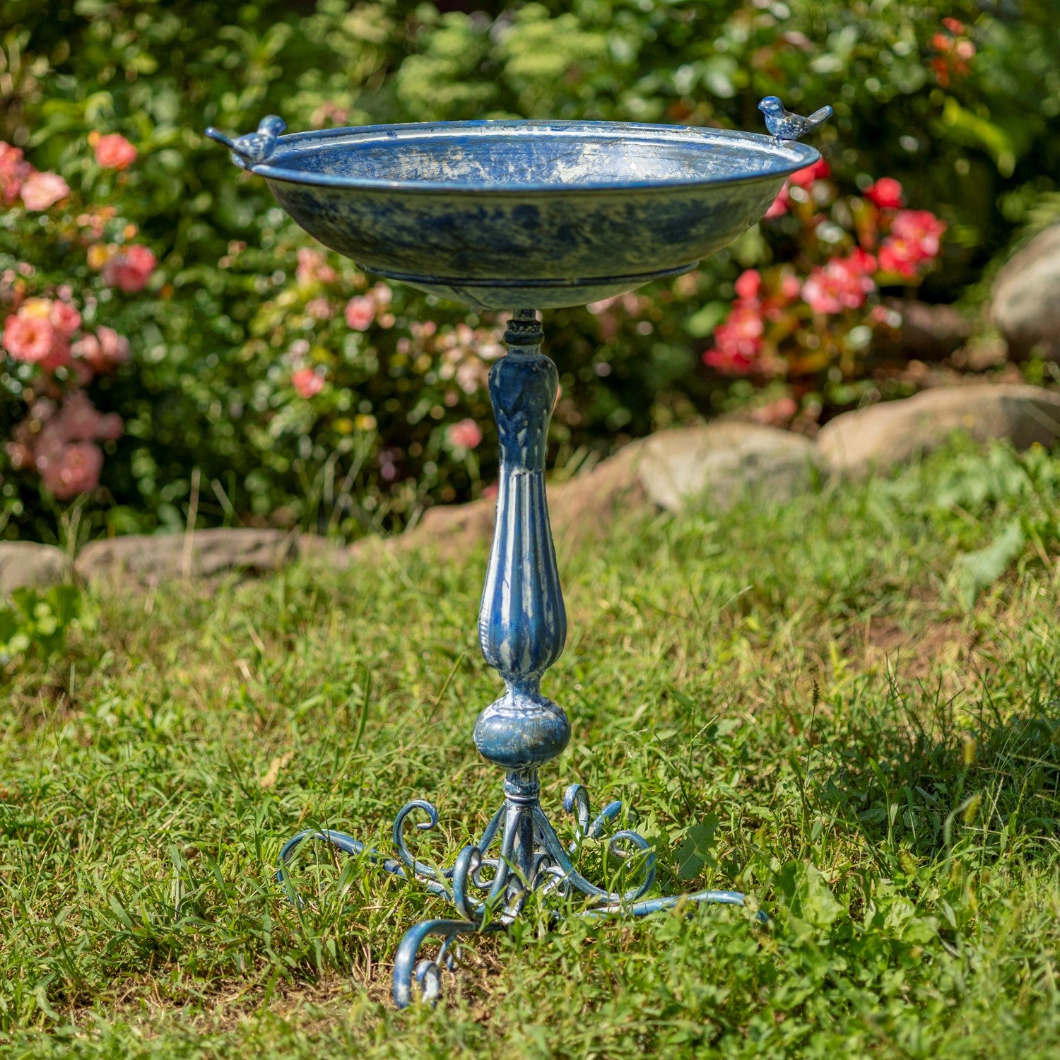 Decorative blue metal bird bath in a garden setting with flowers and rocks.