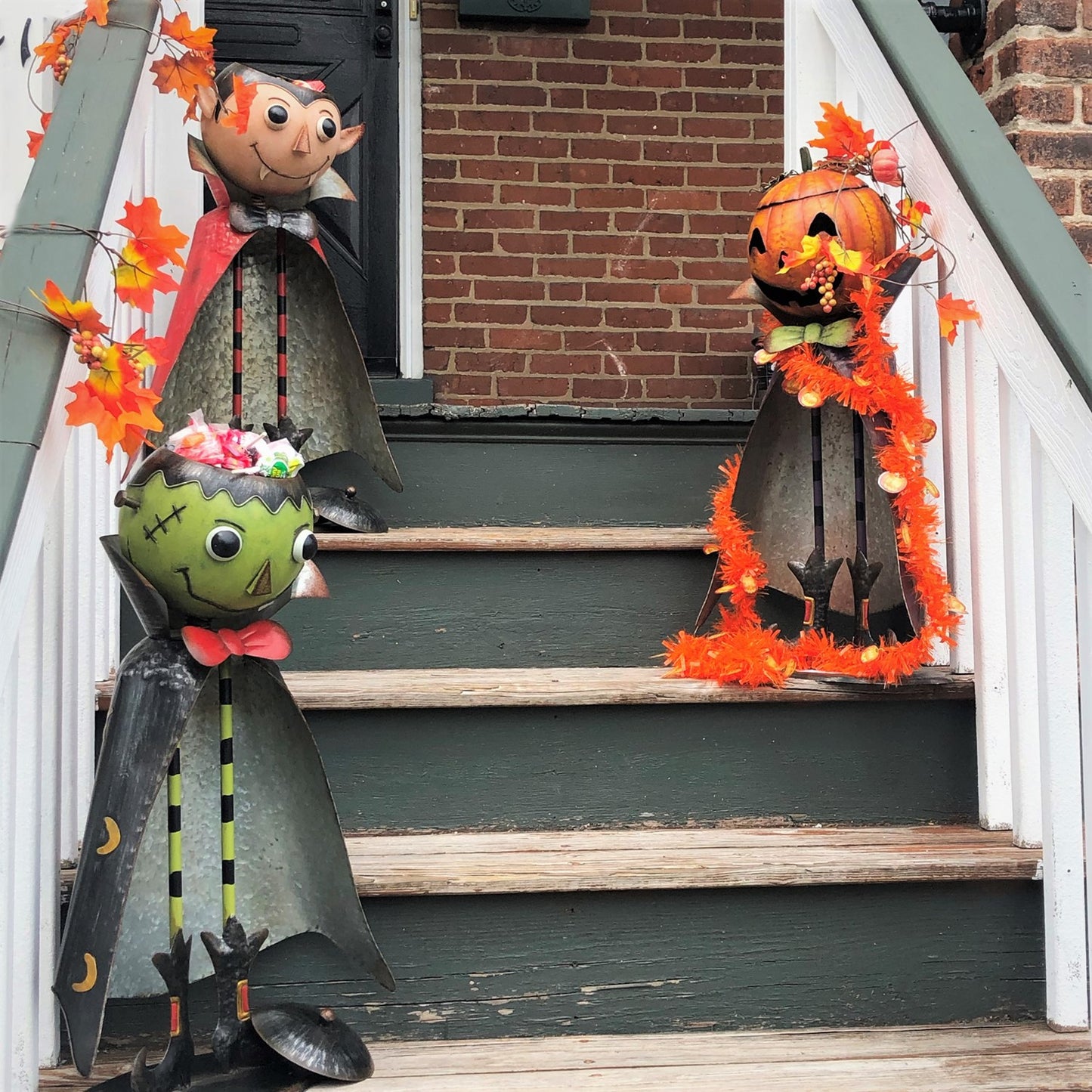 Halloween yard decor on a front porch staircase, featuring a Frankenstein monster, a vampire, and a jack-o'-lantern pumpkin decoration with orange tinsel and autumn leaves.