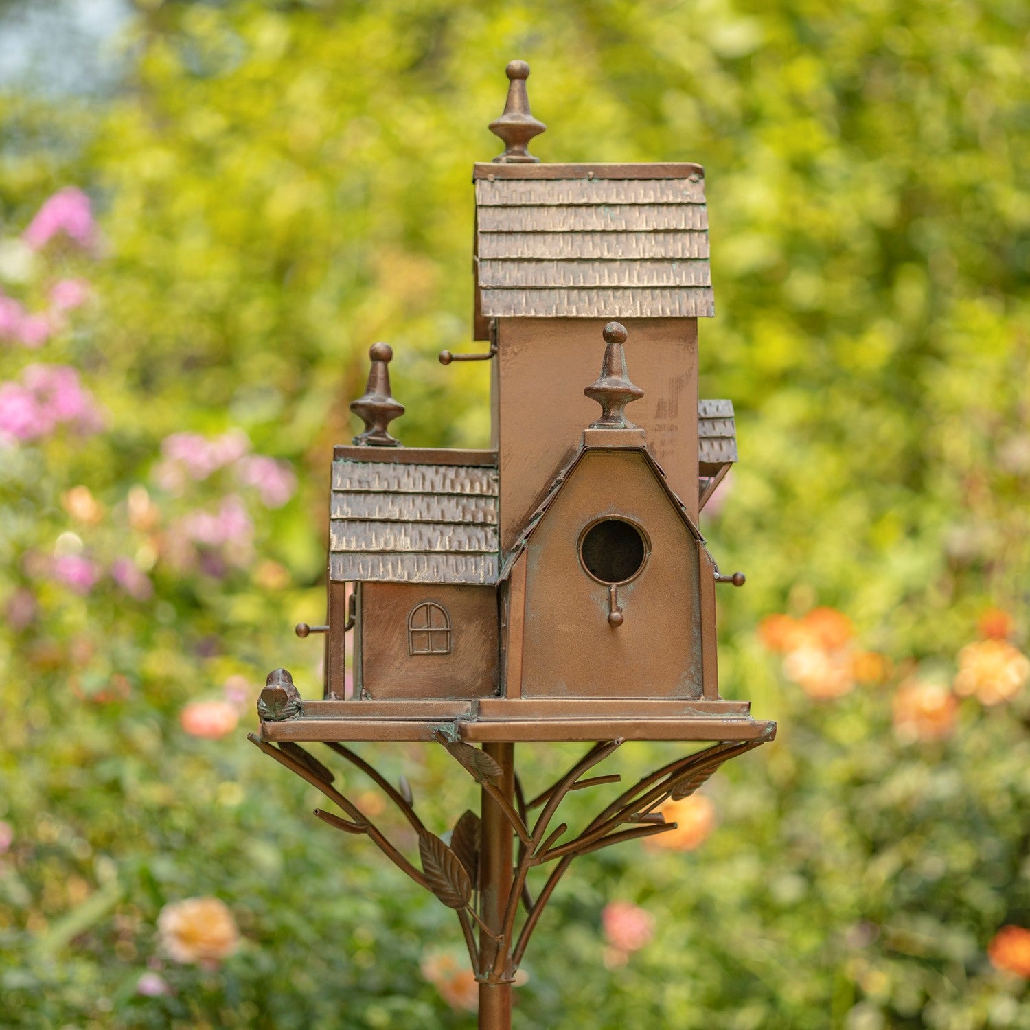 A multi-tiered birdhouse with a rustic brown and bronze finish, mounted on a decorative metal stand with leaf details, set against a blurred green garden background with pink flowers.