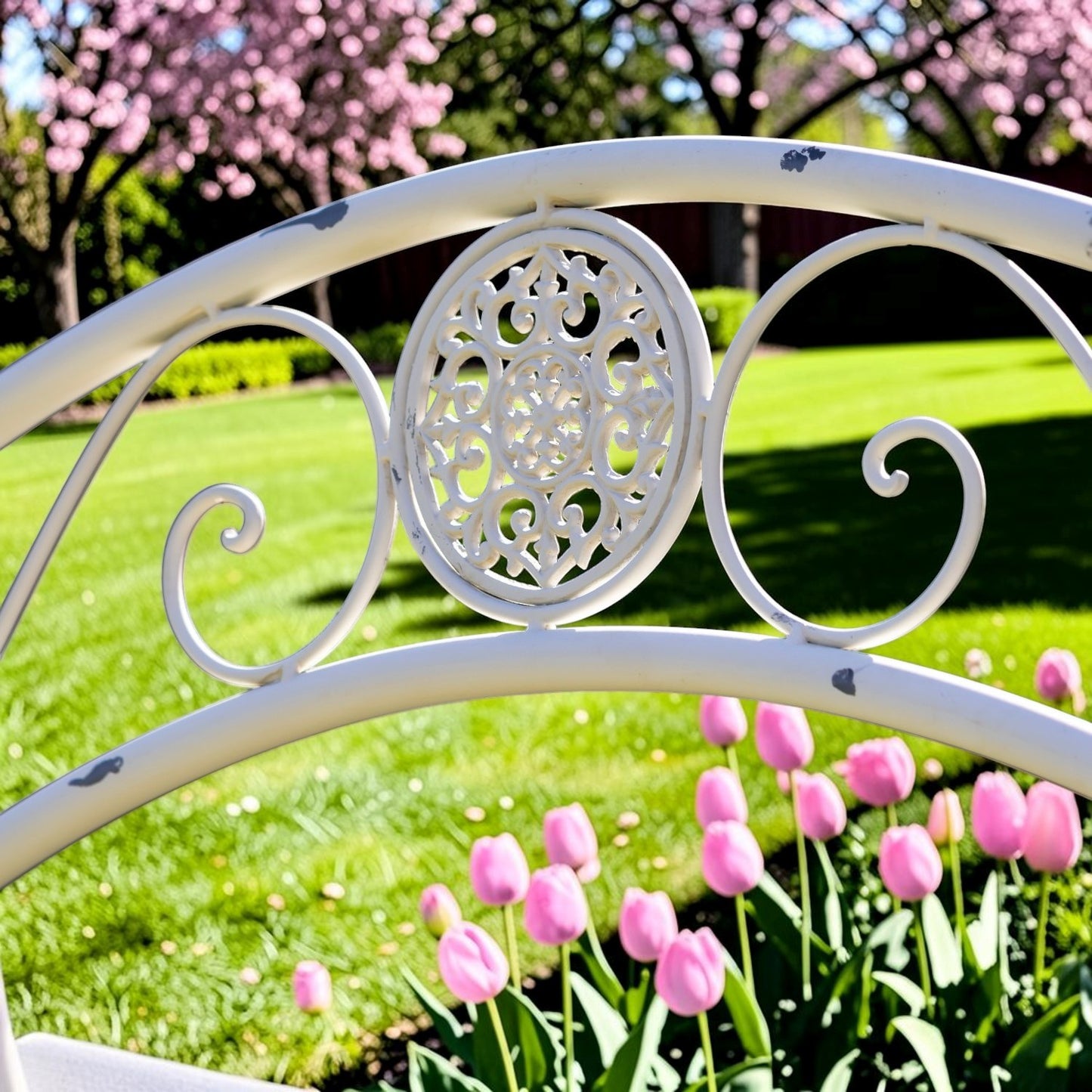 A close-up of a white wrought iron garden bed frame with an ornate central oval cutout, set against a sunny backyard background with pink tulips and cherry blossom trees.