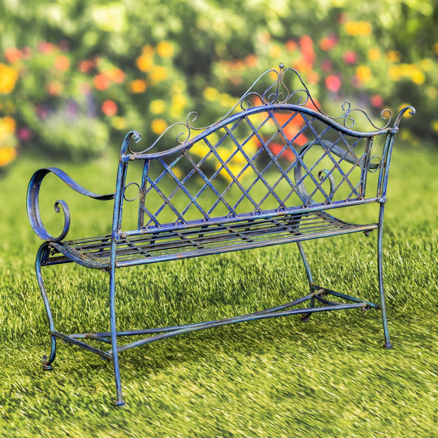 A blue metal garden bench with an ornate diamond-pattern backrest and curved armrests, placed on green grass with a blurred garden background.