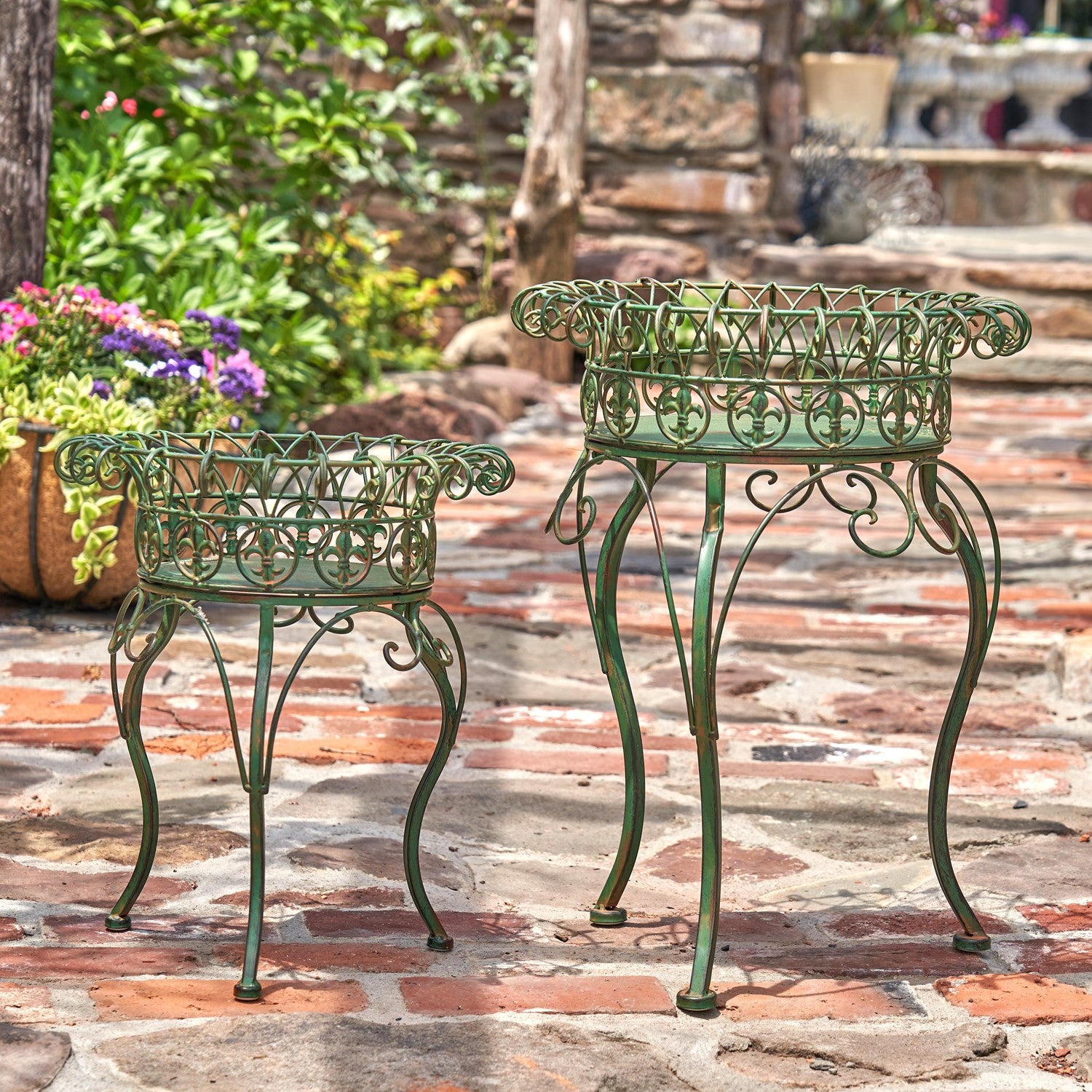 Two green metal garden planters with decorative scrollwork tops and curved legs, displayed on a stone patio in an outdoor garden setting.