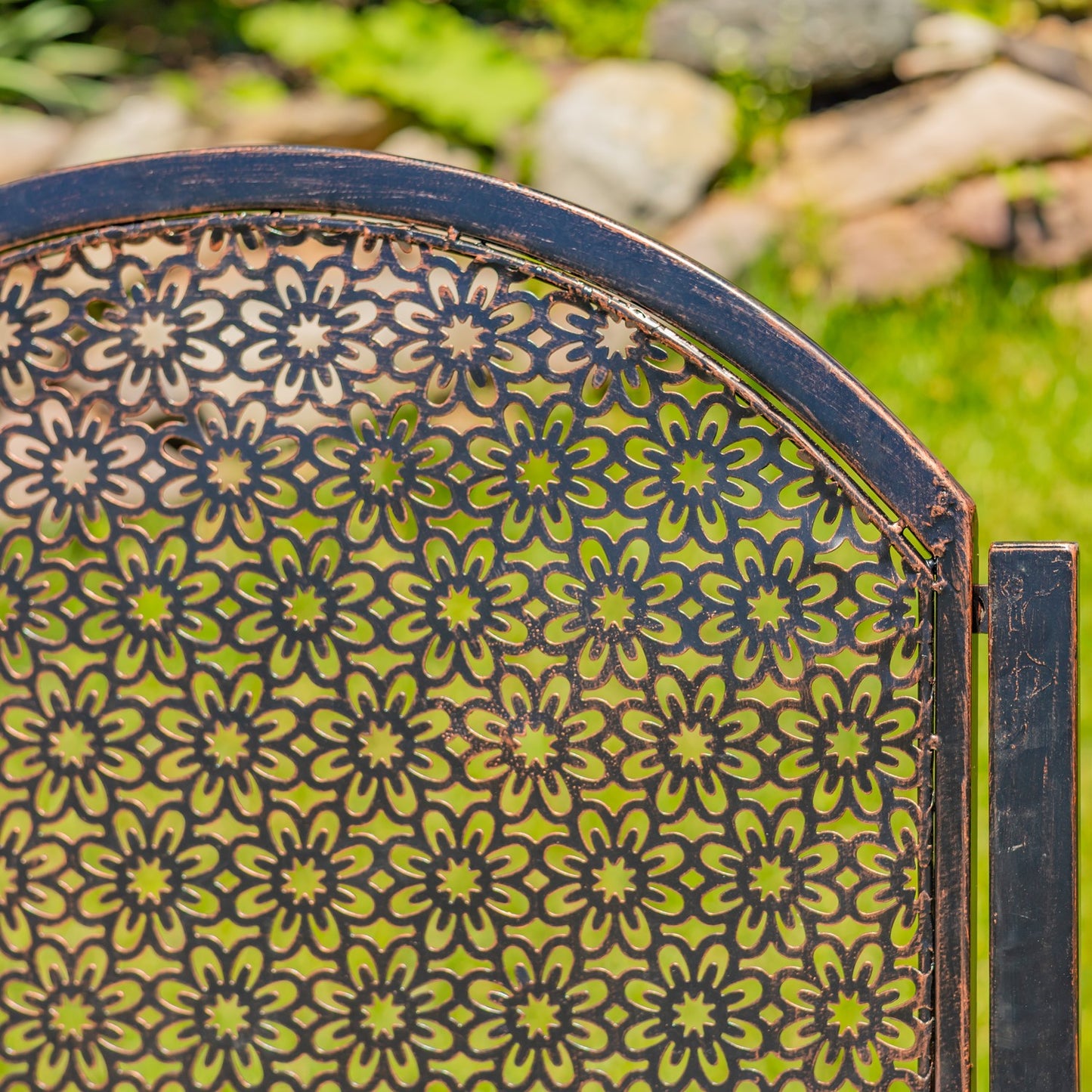 Close-up of a decorative metal garden screen or panel with an arched top and repeating floral cutout pattern, shown outdoors with blurred greenery and rocks in the background.