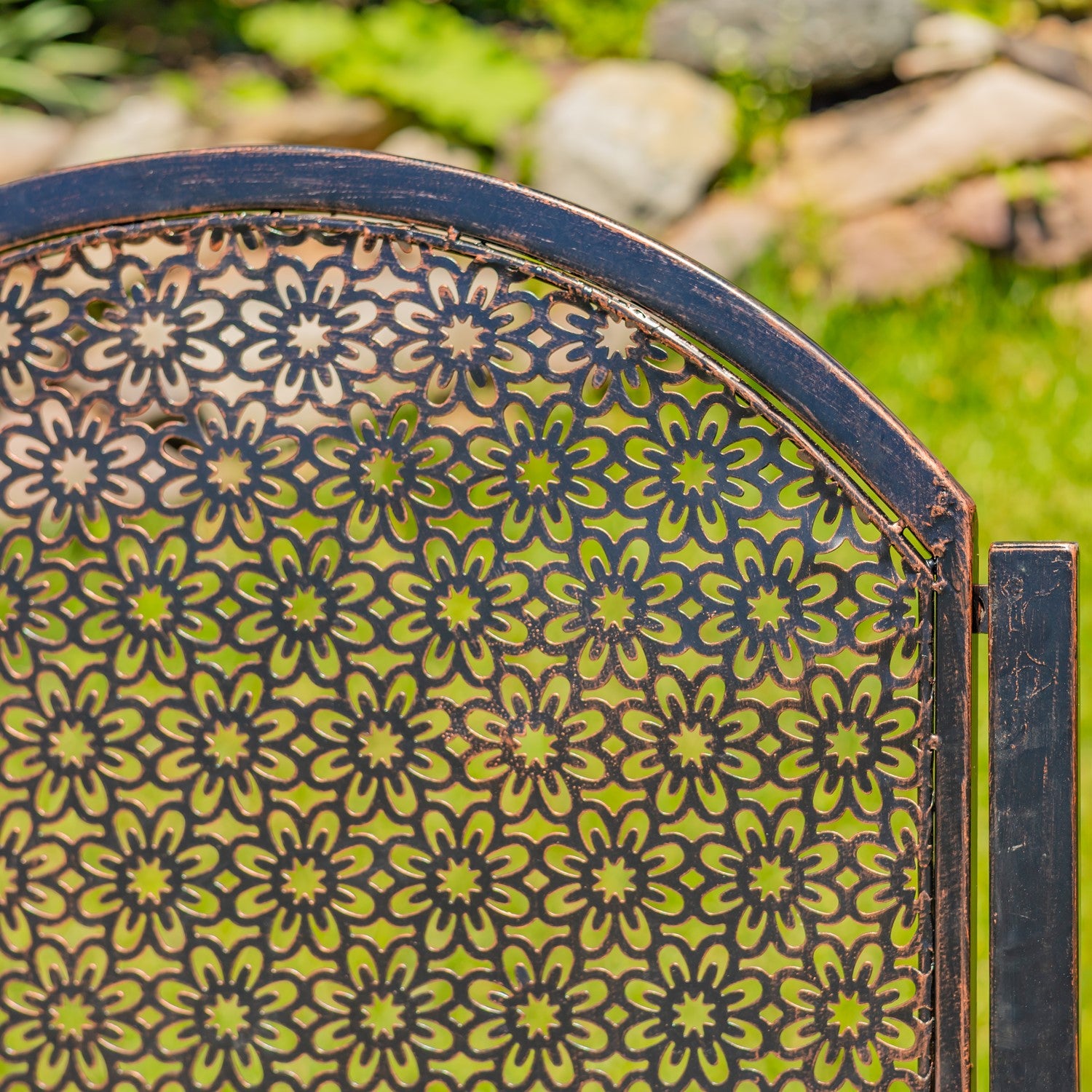 Close-up of a decorative metal garden screen or panel with an arched top and repeating floral cutout pattern, shown outdoors with blurred greenery and rocks in the background.