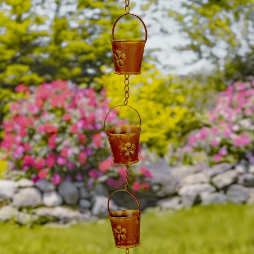 Decorative metal rain chain with floral designs against a garden background with flowers and rocks.