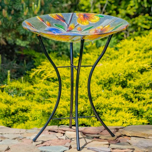 Colorful glass bird bath with floral design on a metal stand outdoors.