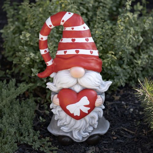 Gnome figurine with a red hat and heart, standing outdoors.