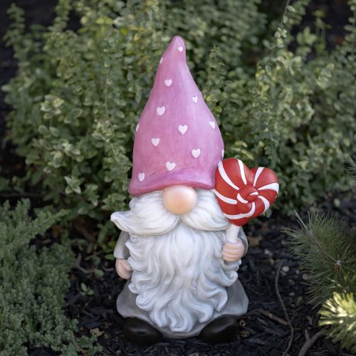 Gnome figurine with a pink hat and white hearts, holding a red heart-shaped lollipop, standing in a garden setting.