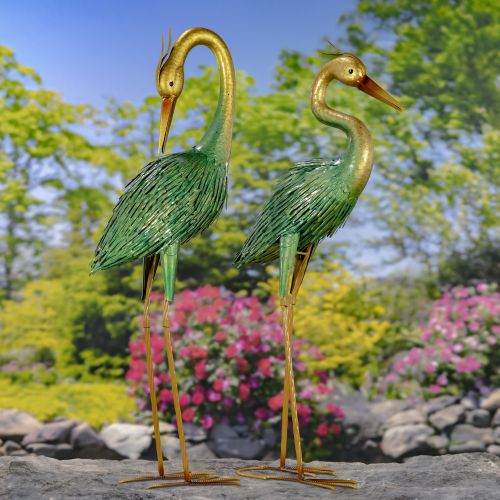 Two green heron statues in a garden setting with flowers and trees.