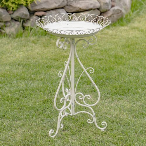 Decorative white metal bird bath on a grassy lawn with a stone wall in the background