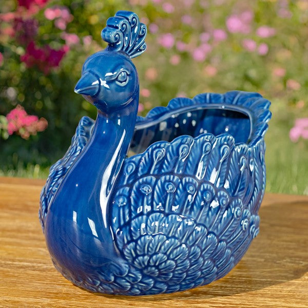 Blue ceramic peacock-shaped planter on a wooden surface with a blurred floral background