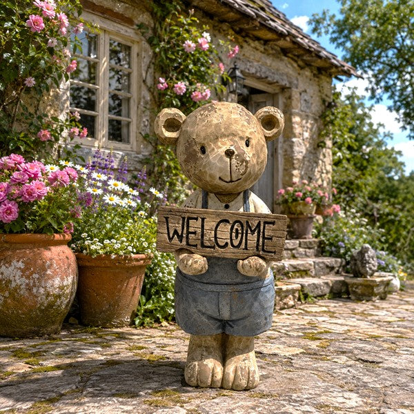 A rustic stone bear garden statue holding a wooden 'WELCOME' sign, set in front of a stone cottage with blooming pink flowers and potted plants.